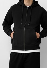 Černá mikina na zip vyrobená z měkkého materiálu, se šňůrkami na kapuci a předními kapsami s vroubkovanými manžetami a lemem. Jednoduchý design.