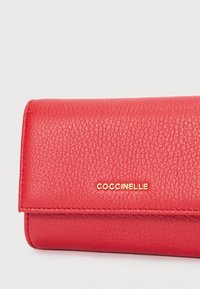 Červená texturovaná kožená peněženka se zlatým logem COCCINELLE umístěným na klopě na jednobarevném světlém pozadí.