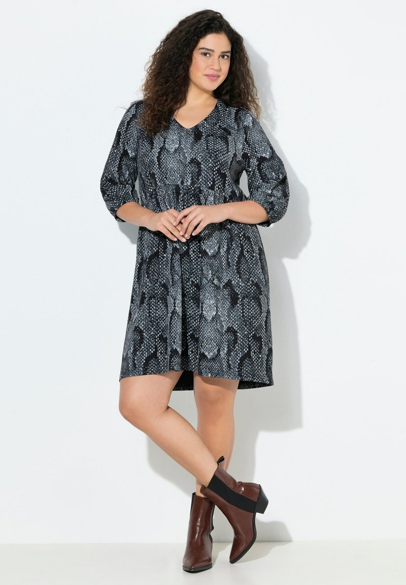 Robe imprimée serpent noir et gris avec des manches trois-quarts, décolleté en V, taille cintrée et jupe évasée. Portée avec des bottines marron à la cheville.