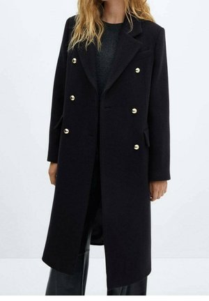 Frau trägt einen langen schwarzen zweireihigen Mantel mit goldenen Knöpfen über einem dunklen Oberteil und schwarzen Hosen vor einem einfarbigen hellen Hintergrund.