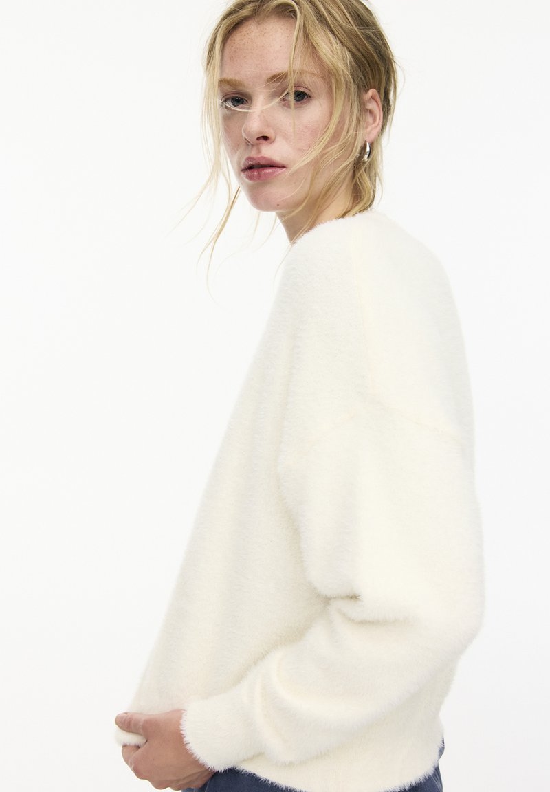 Un pull blanc et duveteux à manches longues, avec un col rond et une texture douce. Vu de côté, avec les mains sur les hanches.
