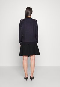 Pull en maille bleu marine avec des poignets et un ourlet côtelés, associé à une jupe noire évasée. La vue arrière montre la couture de l'épaule et une texture lisse.