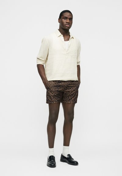 Homme debout portant une chemise à manches longues beige, un short imprimé léopard, des chaussettes blanches et des mocassins noirs sur un fond blanc uni.