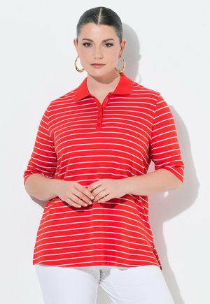 Kvinde med mørkt hår i en rød og hvidstribet polo og hvide bukser, iført store gyldne øreringe, stående foran en hvid baggrund.