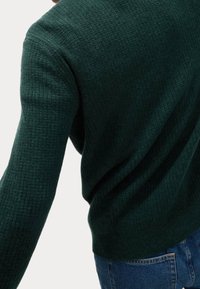 Ciemnozielony teksturowany sweter z długimi rękawami, z subtelnym prążkowanym wzorem i luźnym krojem, połączony z niebieskimi jeansami.