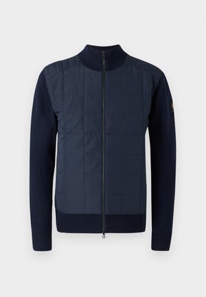 Granatowa kurtka zapinana na zamek, z pikowanym przodem, dzianinowymi rękawami i wysokim kołnierzem. Szczegółowe przeszycia na przednim panelu dodają tekstury.