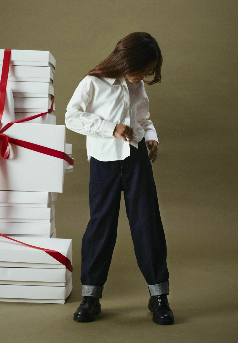 Witte blouse, donkerblauwe wijde jeans met opgerolde manchetten, zwarte schoenen, naast opgestapelde witte cadeau-Dozen met rode linten, neutrale achtergrond.