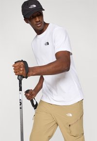 Λευκό βαμβακερό μπλουζάκι με μαύρο λογότυπο, καφέ παντελόνι με φερμουάρ στις τσέπες και μαύρο καπέλο. Κρατάει μπαστούνια πεζοπορίας. Απλή, λειτουργική ένδυση για υπαίθριες δραστηριότητες.