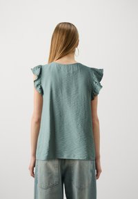 Světlý tyrkysový top s volánovými rukávy, hladká textura, kulatý výstřih a uvolněný střih, v kombinaci se světlými modrými džíny.