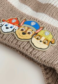 Stickad mössa i beige med ivorifärgade ränder; har tre färgglada hundkaraktärsirepar med hattar, tillverkade av ett slitstarkt gummiliknande material.