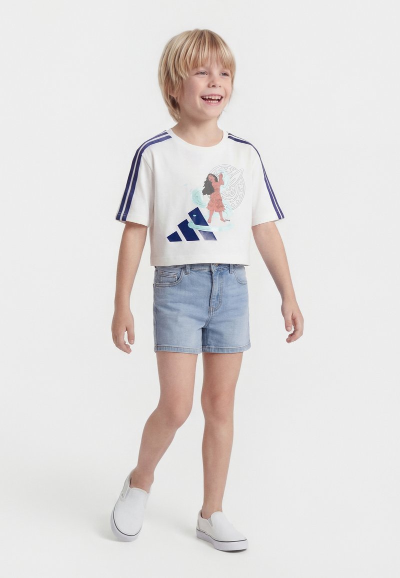 Χαμογελαστό παιδί που φοράει λευκό crop T-shirt με χαρακτήρα κινουμένων σχεδίων, μπλε ρίγες, τζιν σορτς και λευκά παπούτσια slip-on, περπατώντας μπροστά σε λευκό φόντο.