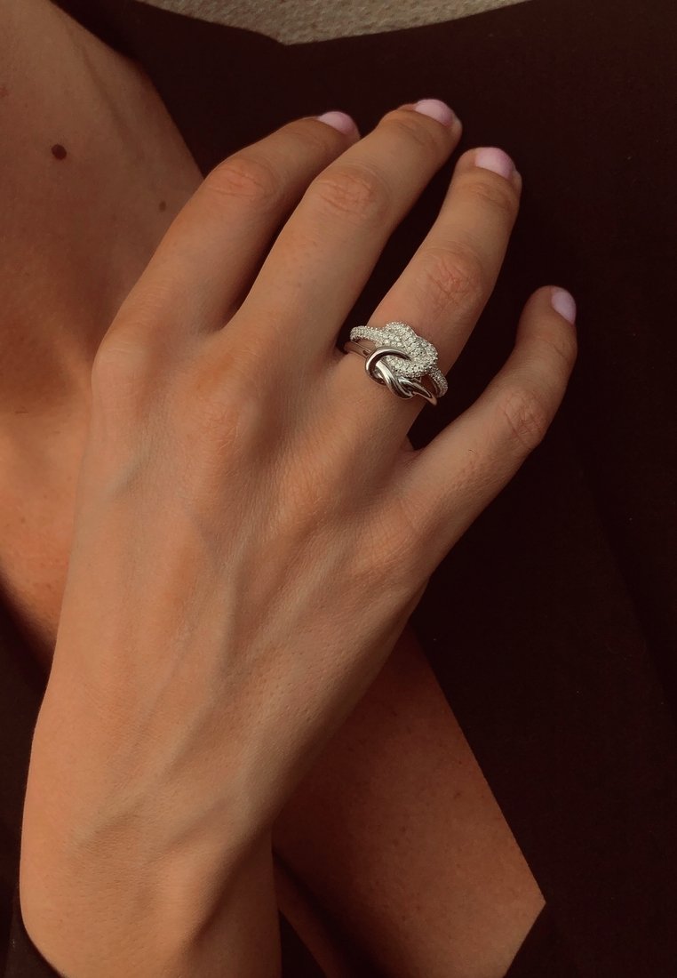 Silberner Ring mit verschlungenem Design, das kleine klare Steine enthält. Hand mit heller Hautfarbe und kurzen pinken Nägeln, die auf einer dunklen Oberfläche ruht.