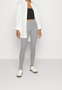Grå leggings gjorda av elastiskt material med en figurnära design, parat med en svart crop top och en oversized vit skjorta. Akkentuerad av en liten logotyp.
