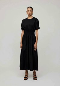 Robe en coton noir à manches courtes, taille smockée et longue jupe jusqu'aux chevilles. Portée avec des sandales noires. Texture lisse, design minimal.