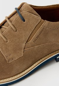 Brauner Wildlederschuh mit einem leicht strukturierten Design, ausgestattet mit blauen Schnürsenkeln und Besatz, geprägtem Logo und einer hellen, genähten Sohle.