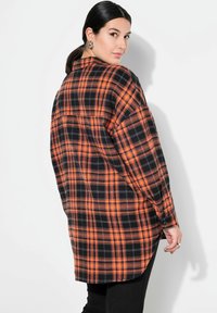 Camicia flanella a quadri nera e arancione con vestibilità rilassata, caratterizzata da maniche lunghe e orlo leggermente curvo. La vista posteriore mostra le spalle scese.