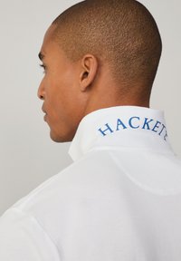 Λευκό πόλο μπλουζάκι με μπλε κεντητό λογότυπο "HACKETT" στο γιακά. Η μπλούζα διαθέτει ανάγλυφο ύφασμα και κομψό σχέδιο.