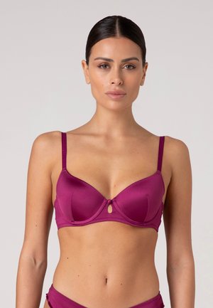 Donna con capelli scuri raccolti in uno chignon liscio che indossa un reggiseno in raso magenta con spalline sottili e un piccolo taglio frontale, su uno sfondo chiaro e uniforme.