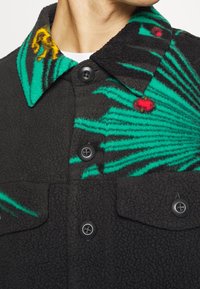 Czarna polarowa kurtka z zielonym i żółtym wzorem liści palmowych. Posiada czarne guziki i teksturowany kołnierz. Zawiera kieszeń na piersi.