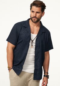 Dunkelblaues kurzärmliges Hemd mit strukturierten geometrischen Mustern, Button-Down-Kragen und lockerer Passform, über einem weißen Tanktop getragen.