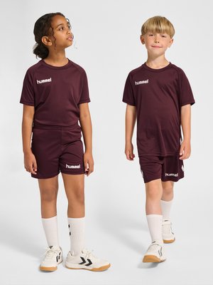 Zwei Kinder in passenden bordeauxroten Sportkleidungen und weißen Sneakers stehen vor einem schlichten Hintergrund, eines schaut seitlich und das andere geradeaus.