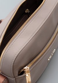 Borsa beige con due cerniere dorate visibili, una parzialmente aperta che mostra una tasca interna in tessuto scuro con una tasca aggiuntiva con cerniera.