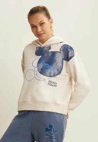 Felpa crema con grafica di Mickey Mouse blu; tessuto morbido; tasca frontale; abbinata a pantaloni joggers blu navy con logo coordinato.