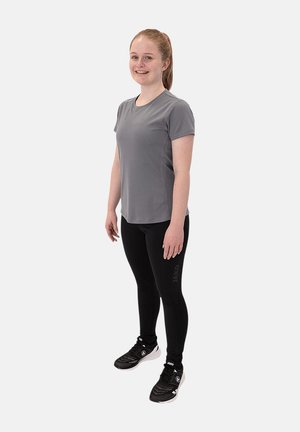 Jeune femme debout et souriante, portant un t-shirt gris à manches courtes, des leggings noirs et des chaussures de sport noires.