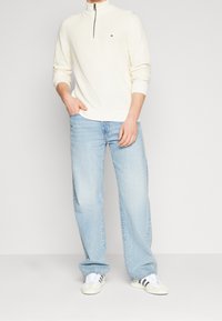 Muž oblečený v krémovém texturovaném svetr s polo zipem, světle modrých volných džínách a bílých teniskách s černými pruhy, stojící na světlém pozadí.