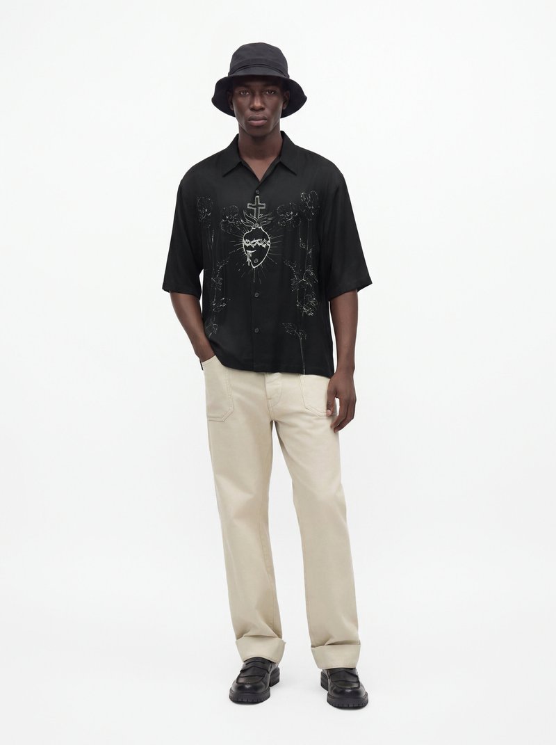 Homme portant un chapeau bob noir, une chemise noire avec un motif croix et cœur, un pantalon beige et des chaussures noires, debout sur un fond blanc.