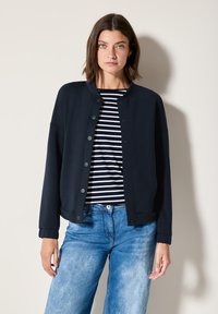 Giacca leggera blu navy con bottoni a pressione, colletto tondo e maniche lunghe, indossata sopra una maglietta a righe e jeans blu.