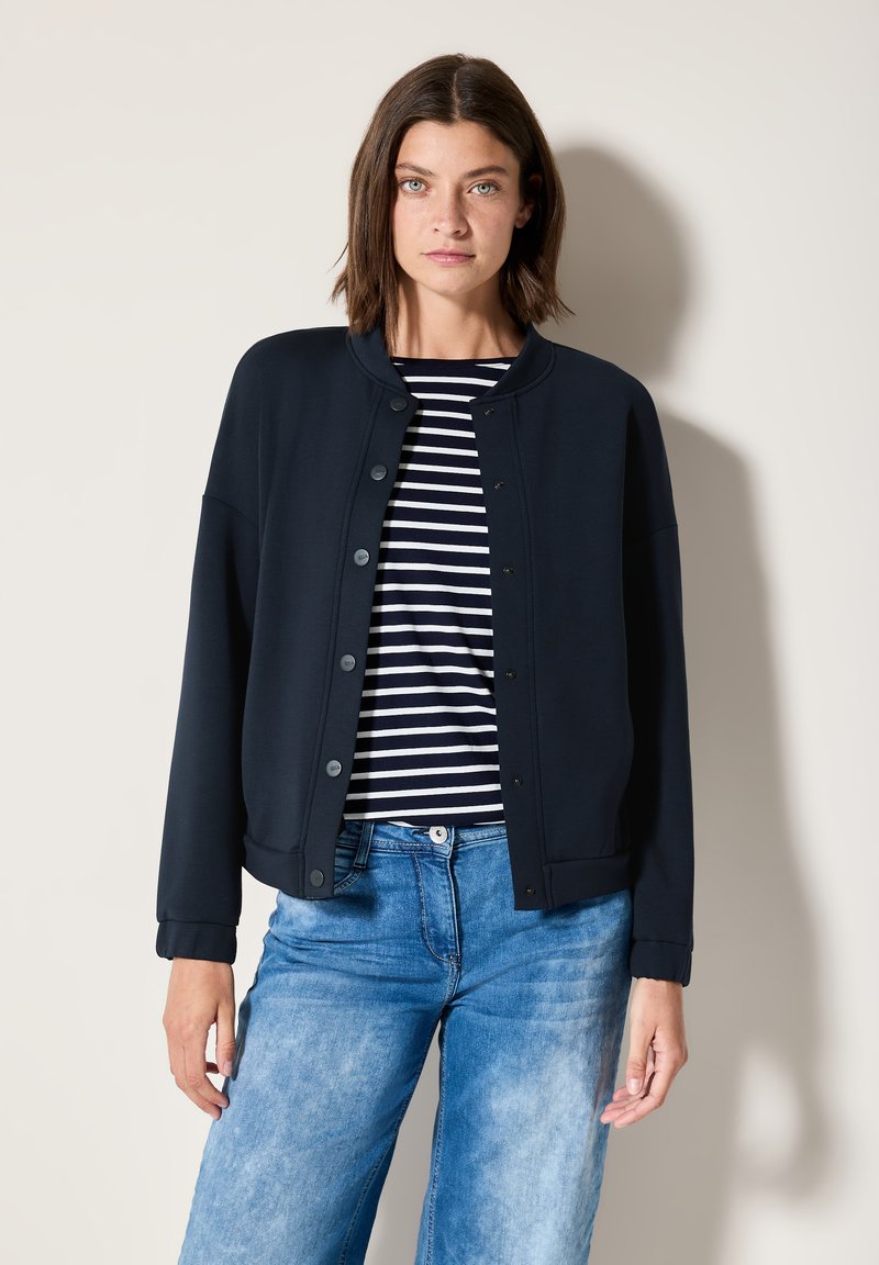 Giacca leggera blu navy con bottoni a pressione, colletto tondo e maniche lunghe, indossata sopra una maglietta a righe e jeans blu.