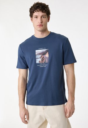 Młody mężczyzna o kręconych brązowych włosach, ubrany w granatowy t-shirt z grafiką nadmorskiej drogi oraz beżowe spodnie, na białym tle.