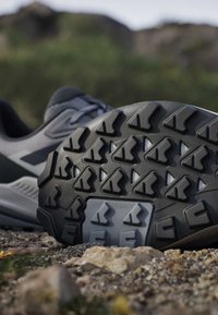 Close-up of a gray and black trail running shoe showing its textured, rugged sole on rocky outdoor ground.