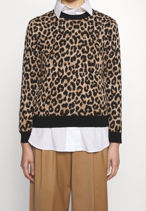 Personne portant un pull à motif léopard sur une chemise blanche à col, associée à un pantalon marron ample, debout devant un fond uni.