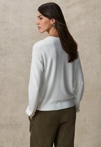 Witte gebreide trui met een geribbelde zoom, ronde hals en verlaagde schouders, gecombineerd met olijfgroene broek. Model heeft lang haar en is gezien vanuit de rug.