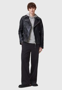Chaqueta de motociclista de cuero negra con cremallera en el frente y bolsillos, superpuesta a una sudadera gris con capucha, combinada con pantalones sueltos negros y zapatillas.
