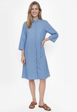 Vrouw met een glimlach staat met één hand op haar heup, draagt een blauw overhemdjurk tot aan de knie en bruine sandalen op een witte achtergrond.