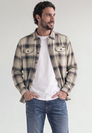 Homme portant une chemise à carreaux beige et noire sur un t-shirt blanc avec un jean bleu, les mains dans les poches, regardant sur le côté, fond neutre.