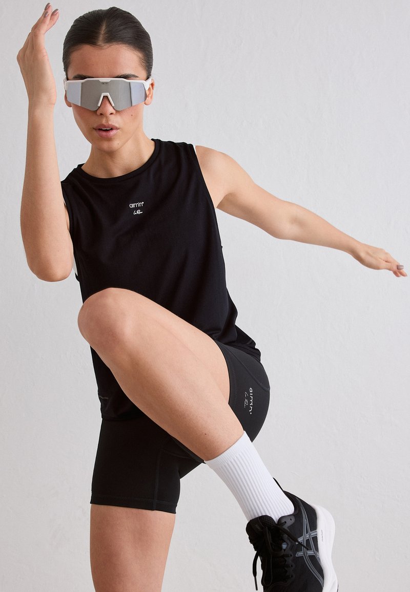 Femme en tenue de sport noire et lunettes de soleil réfléchissantes, levant un genou et tendant le bras sur un fond clair uni.