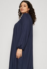 Tamsiai mėlyna, ilgomis rankovėmis su laisvu siluetu ir pūkuotomis rankovėmis, pagaminta iš minkštos medžiagos. Modelis stovi šoniniame vaizde.