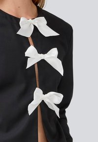 Svart jakke med tre hvite satengsløyfer som lukking foran. Glatt tekstur og minimalistisk design med en subtil åpning.