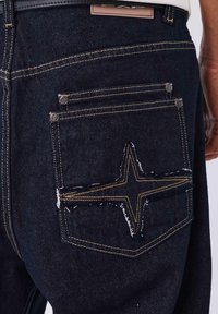 Los vaqueros de denim presentan un lavado oscuro con costuras amarillas. El bolsillo trasero tiene un diseño distintivo de estrella con bordes deshilachados y detalles de herrajes plateados.