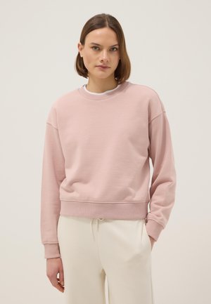 Vrouw met halflang bruin haar, gekleed in een lichtroze sweatshirt en crème-kleurige broek, staand tegen een effen achtergrond.