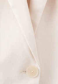 Prim-plan al unui sacou din in bej, cu rever cu creastă și un singur nasture bej deschis în față.