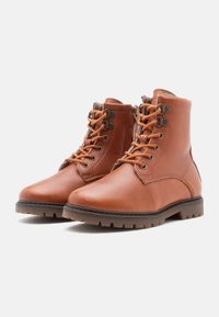 Bottes de cheville en cuir marron à lacets avec fermeture éclair sur le côté, intérieur rembourré et semelles en caoutchouc épaisses, légèrement inclinées à droite sur un fond blanc.