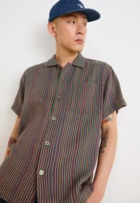Short-sleeve button-up shirt featuring a multicolored striped pattern, lightweight fabric, and a chest pocket. Pairing with a blue cap.