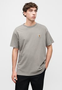 Γκρι βαμβακερό t-shirt με κοντά μανίκια, στρογγυλή λαιμόκοψη και τετράγωνο λογότυπο στο αριστερό στήθος. Φορεμένο με σκούρο παντελόνι.