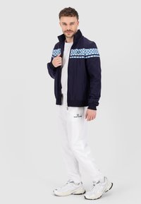 Giacca bomber navy con righe a quadri blu, t-shirt bianca, joggers bianchi e sneakers sportive. Caratterizzata da tasche laterali e chiusura con zip.