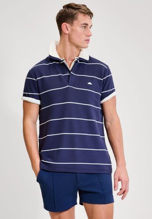 Mężczyzna w granatowej koszulce polo z białymi poziomymi paskami i dopasowanych granatowych szortach, patrzący na bok.